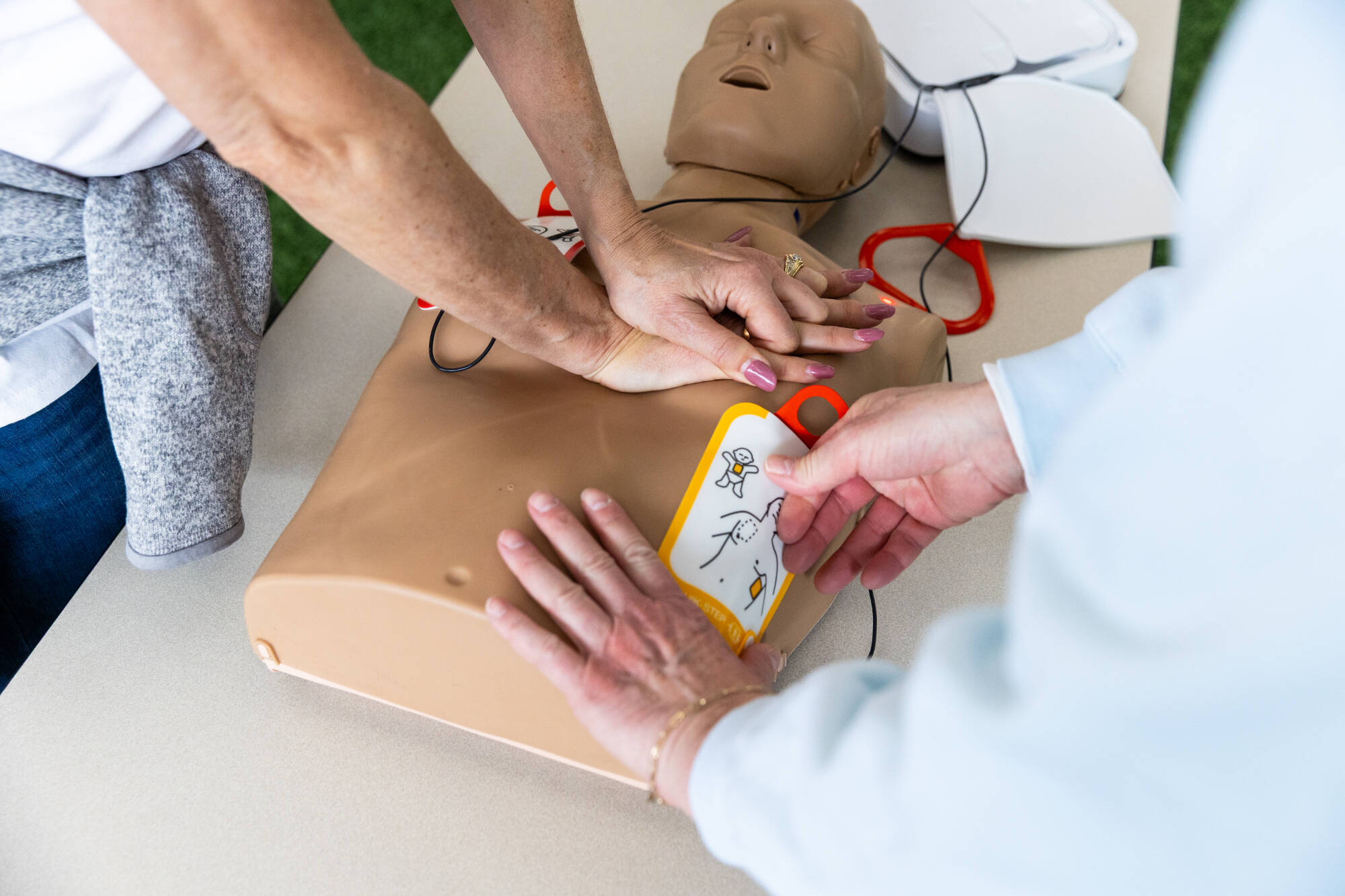 An instructor performs chest compressions as volunteer places AED pads during a CPR training session.